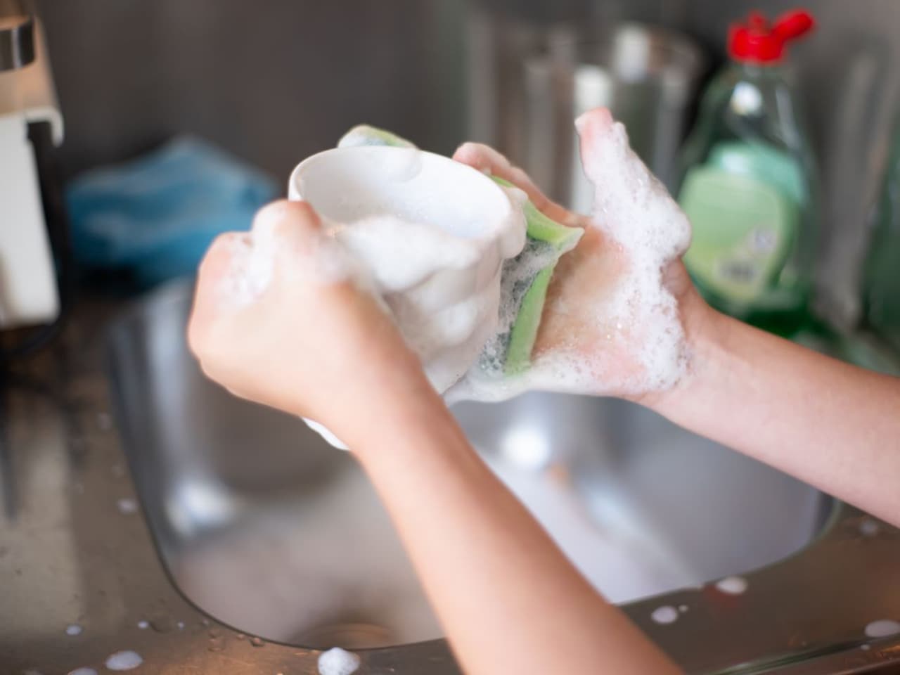 How to clean an oven with baking soda and water: hands scrubbing with a soapy sponge over a steel sink